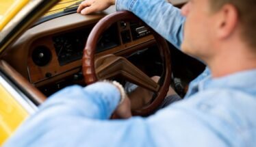 Driver in a blue shirt gripping the brown steering wheel inside a yellow vintage car interior.