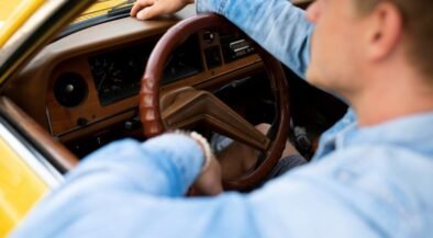 Driver in a blue shirt gripping the brown steering wheel inside a yellow vintage car interior.