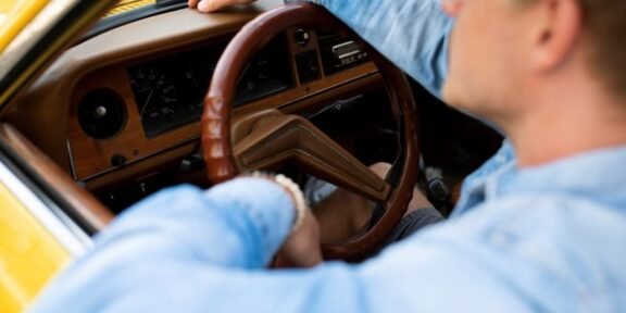 Driver in a blue shirt gripping the brown steering wheel inside a yellow vintage car interior.
