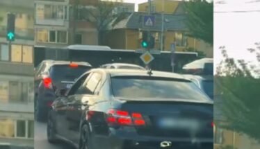 Rear view of a dark sedan in a traffic jam on a city street with apartment buildings and trees in the background.