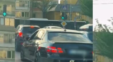Rear view of a dark sedan in a traffic jam on a city street with apartment buildings and trees in the background.
