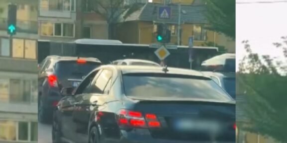 Rear view of a dark sedan in a traffic jam on a city street with apartment buildings and trees in the background.