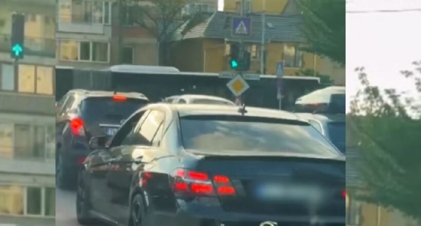 Rear view of a dark sedan in a traffic jam on a city street with apartment buildings and trees in the background.