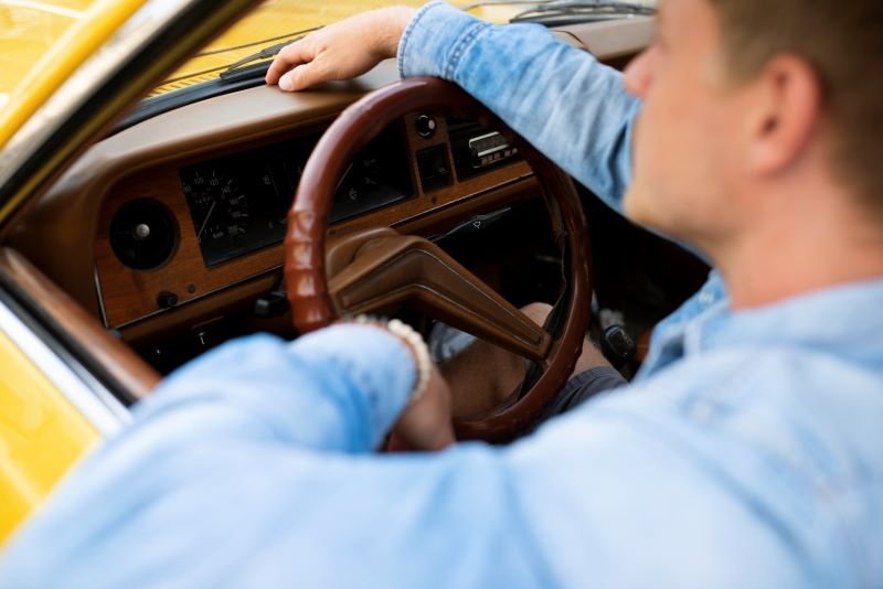 Driver in a blue shirt gripping the brown steering wheel inside a yellow vintage car interior.