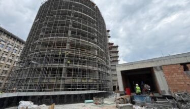 Cylindrical building under heavy scaffolding at an active construction site, with workers near a tunnel-like entrance nearby.
