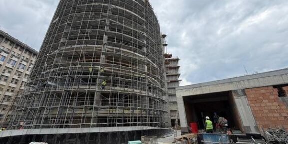 Cylindrical building under heavy scaffolding at an active construction site, with workers near a tunnel-like entrance nearby.
