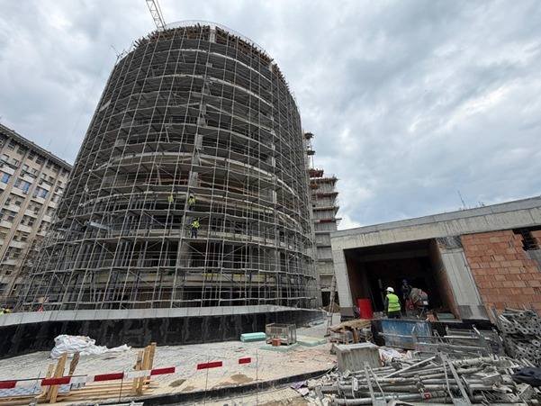 Cylindrical building under heavy scaffolding at an active construction site, with workers near a tunnel-like entrance nearby.