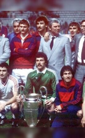 Group of soccer players and coaches posing with a large trophy on a grass field, team members kneeling in front and others standing behind.