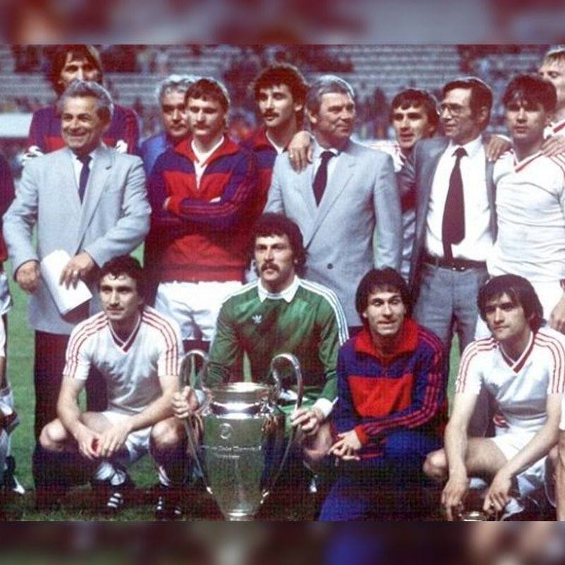 Group of soccer players and coaches posing with a large trophy on a grass field, team members kneeling in front and others standing behind.