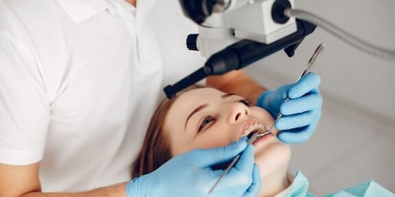 Dentist in blue gloves examines a patient’s teeth under a dental microscope.