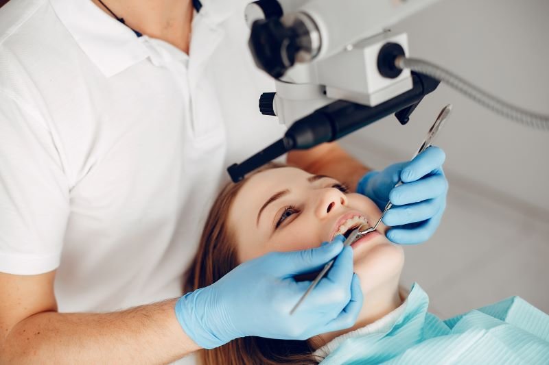 Dentist in blue gloves examines a patient’s teeth under a dental microscope.