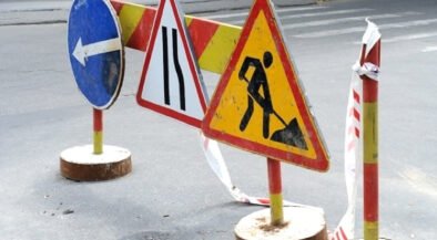 Road work zone with warning signs and barriers on an asphalt street; includes a blue left-turn sign, lane merge sign, and digging worker symbol with tape barriers