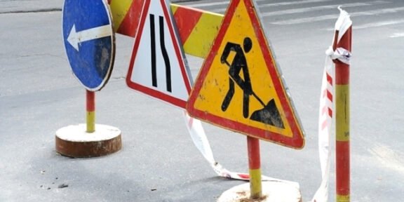 Road work zone with warning signs and barriers on an asphalt street; includes a blue left-turn sign, lane merge sign, and digging worker symbol with tape barriers
