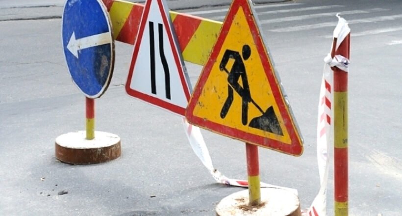 Road work zone with warning signs and barriers on an asphalt street; includes a blue left-turn sign, lane merge sign, and digging worker symbol with tape barriers