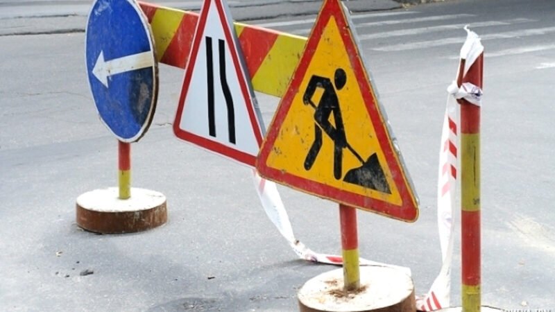 Road work zone with warning signs and barriers on an asphalt street; includes a blue left-turn sign, lane merge sign, and digging worker symbol with tape barriers