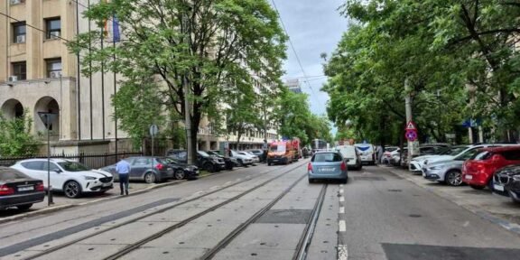 City street with tram tracks down the middle, parked cars on both sides, and a beige building on the left with green trees lining the sidewalk.