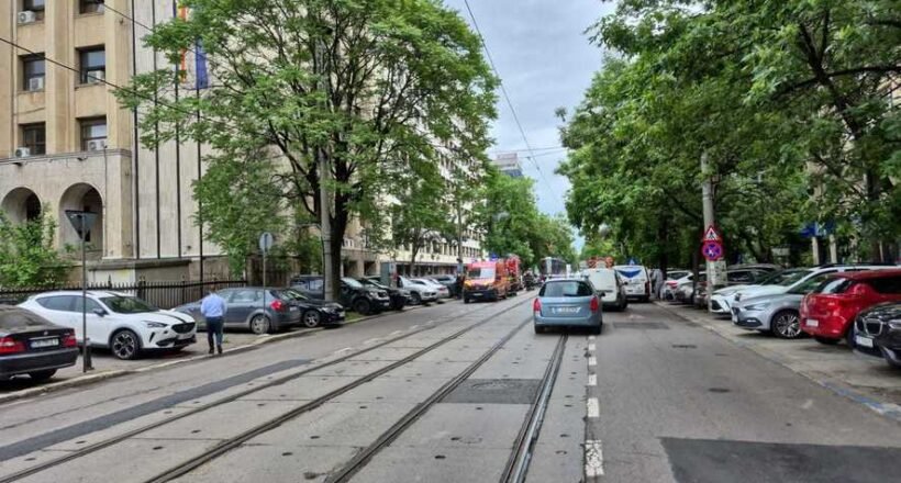 City street with tram tracks down the middle, parked cars on both sides, and a beige building on the left with green trees lining the sidewalk.