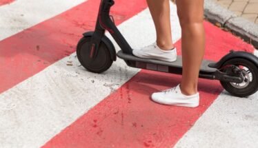 Person wearing white sneakers standing on a black electric scooter on a red-and-white crosswalk pavement.