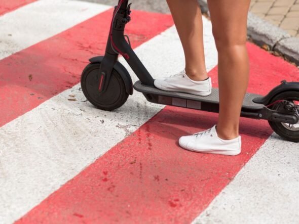 Person wearing white sneakers standing on a black electric scooter on a red-and-white crosswalk pavement.
