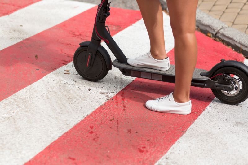 Person wearing white sneakers standing on a black electric scooter on a red-and-white crosswalk pavement.