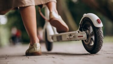 Person wearing beige pants and sneakers stands next to a white kick scooter on a paved path, viewed low from the ground.