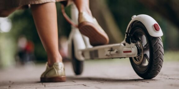 Person wearing beige pants and sneakers stands next to a white kick scooter on a paved path, viewed low from the ground.