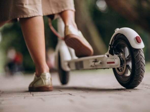 Person wearing beige pants and sneakers stands next to a white kick scooter on a paved path, viewed low from the ground.