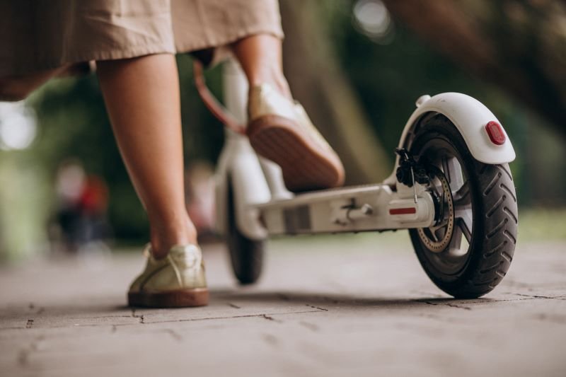 Person wearing beige pants and sneakers stands next to a white kick scooter on a paved path, viewed low from the ground.