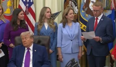 Man in a dark suit and magenta tie sits at a desk while officials stand behind him in a formal ceremony with flags in the background.