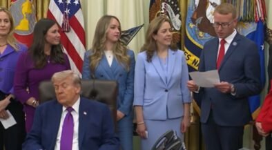 Man in a dark suit and magenta tie sits at a desk while officials stand behind him in a formal ceremony with flags in the background.