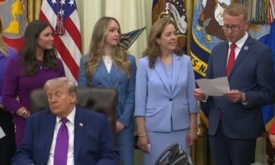 Man in a dark suit and magenta tie sits at a desk while officials stand behind him in a formal ceremony with flags in the background.