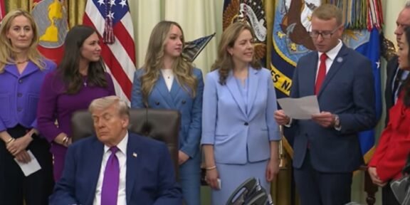 Man in a dark suit and magenta tie sits at a desk while officials stand behind him in a formal ceremony with flags in the background.