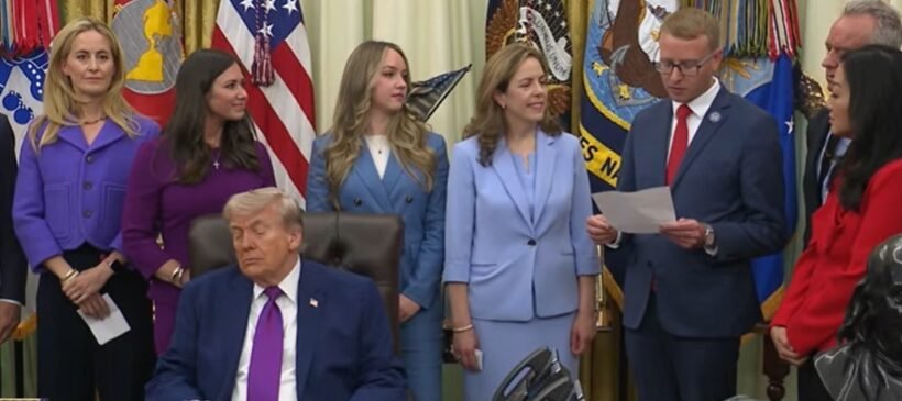 Man in a dark suit and magenta tie sits at a desk while officials stand behind him in a formal ceremony with flags in the background.