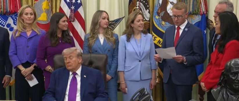 Man in a dark suit and magenta tie sits at a desk while officials stand behind him in a formal ceremony with flags in the background.