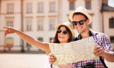 Couple wearing sun hats and sunglasses looking at a map while exploring a sunny city square; one points into the distance.