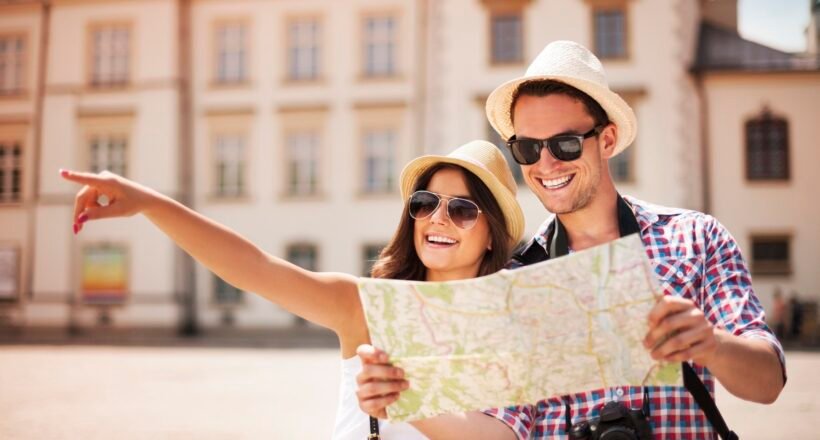 Couple wearing sun hats and sunglasses looking at a map while exploring a sunny city square; one points into the distance.
