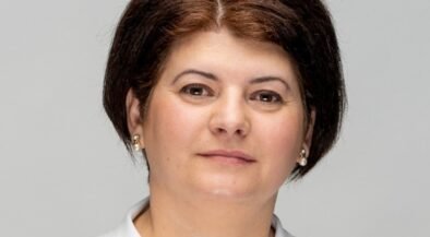 Professional head-and-shoulders portrait of a woman with short dark hair, pearl earrings, and a light gray background.