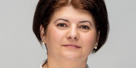 Professional head-and-shoulders portrait of a woman with short dark hair, pearl earrings, and a light gray background.