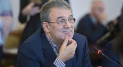 Smiling middle-aged man with gray hair and glasses in a blue suit, seated at a conference table with a microphone nearby, resting his chin on his hand.