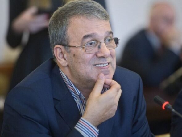 Smiling middle-aged man with gray hair and glasses in a blue suit, seated at a conference table with a microphone nearby, resting his chin on his hand.
