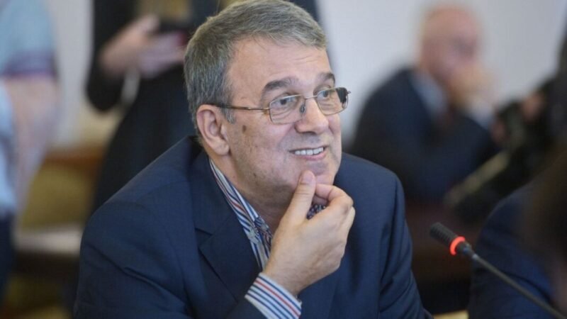 Smiling middle-aged man with gray hair and glasses in a blue suit, seated at a conference table with a microphone nearby, resting his chin on his hand.