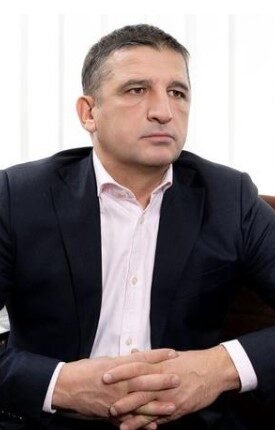 Man in a navy suit and pink shirt sits at a desk with hands clasped, in an office with blinds and a plant in the background.