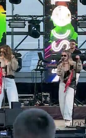 A live outdoor band performing on stage with two female vocalists, a drummer, a guitarist, and bright screens behind them.
