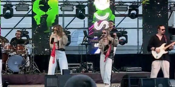 A live outdoor band performing on stage with two female vocalists, a drummer, a guitarist, and bright screens behind them.