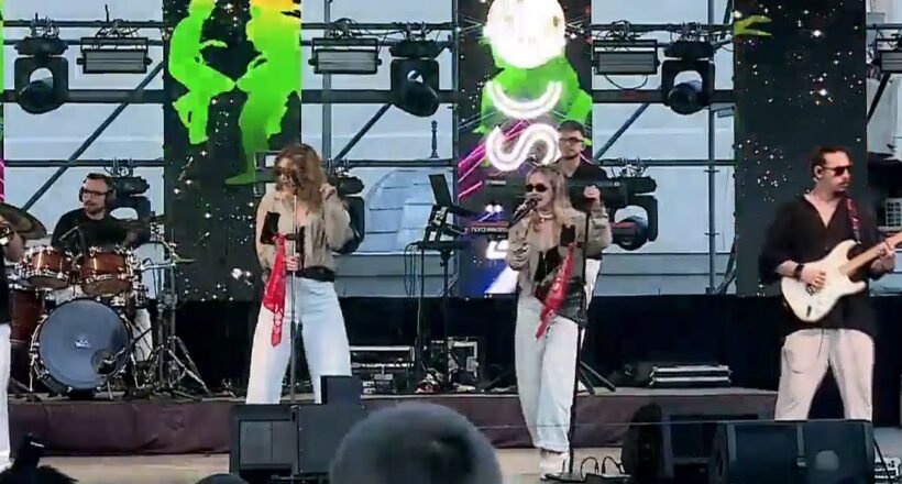 A live outdoor band performing on stage with two female vocalists, a drummer, a guitarist, and bright screens behind them.