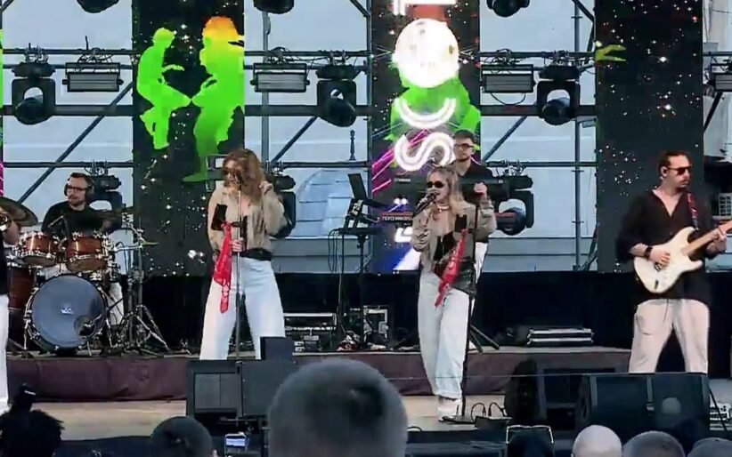 A live outdoor band performing on stage with two female vocalists, a drummer, a guitarist, and bright screens behind them.