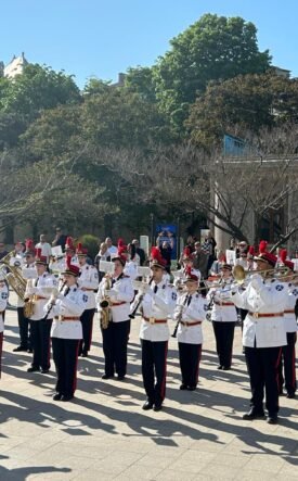 Military band in white uniforms performing a parade in a sunlit square, with a Greek flag on the right.