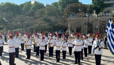 Military band in white uniforms performing a parade in a sunlit square, with a Greek flag on the right.