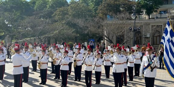 Military band in white uniforms performing a parade in a sunlit square, with a Greek flag on the right.