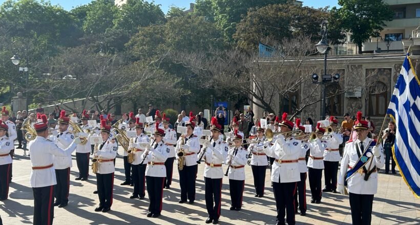 Military band in white uniforms performing a parade in a sunlit square, with a Greek flag on the right.